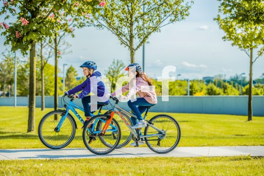 Zwei Kinder fahren auf Fahrrädern einen Betonweg zwischen Rasenflächen entlang.