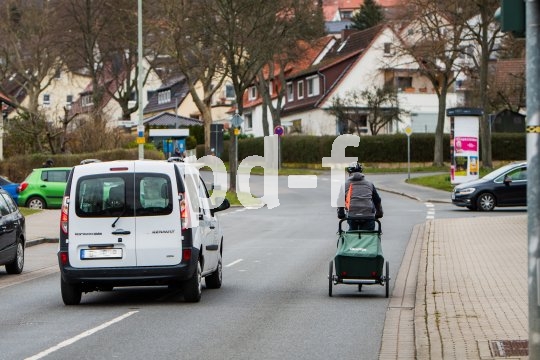Ein weißes Auto überholt eine Person auf einem Fahrrad mit Anhänger auf einer Straße ohne Fahrradspur in einem Wohngebiet.