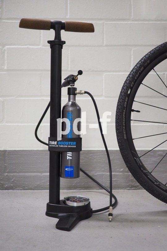 Neben einem Rad steht eine Fahrrad-Standpumpe mit einer Metallflasche und mehrere Schläuchen daran.