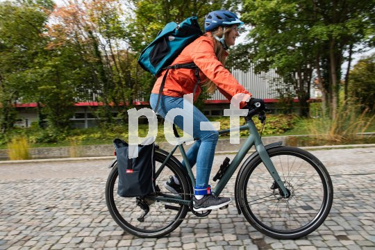 Eine Radfahrerin mit Rucksack und Gepäcktasche am Fahrrad fährt eine Straße mit Kopfsteinpflaster entlang.