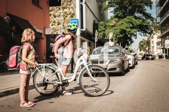 Ein Mädchen mit und eins ohne Fahrrad stehen am Straßenrand und schauen nach Autos, bevor sie die Straße überqueren.