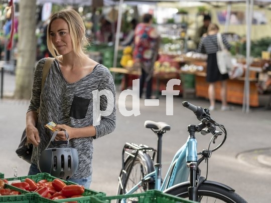 Eine Frau steht mit Fahrradhelm in der Hand an einem Gemüsestand auf einem Markt. Neben ihr steht ein E-Bike.