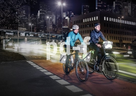 Zwei Personen auf Fahrrädern mit Licht und reflektierenden Elementen an den Fahrradtaschen vor vielen Lichtern einer Großstadt.