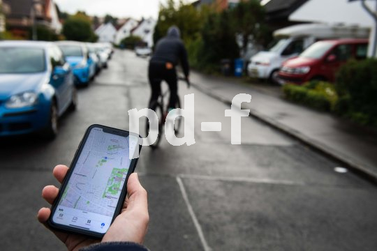 Eine Hand mit einem Smartphone, das einen Stadtplan anzeigt. Unscharf im Hintergrund ist eine Person auf einem Fahrrad in einem Wohngebiet zu sehen.