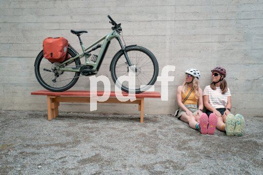 Ein E-Bike mit roten Taschen am Gepäckträger steht auf einer Bank an eine Wand gelehnt. Neben der Bank sitzen zwei Frauen mit Fahrradhelmen auf dem Boden an der Wand und schauen in Richtung des Rades.