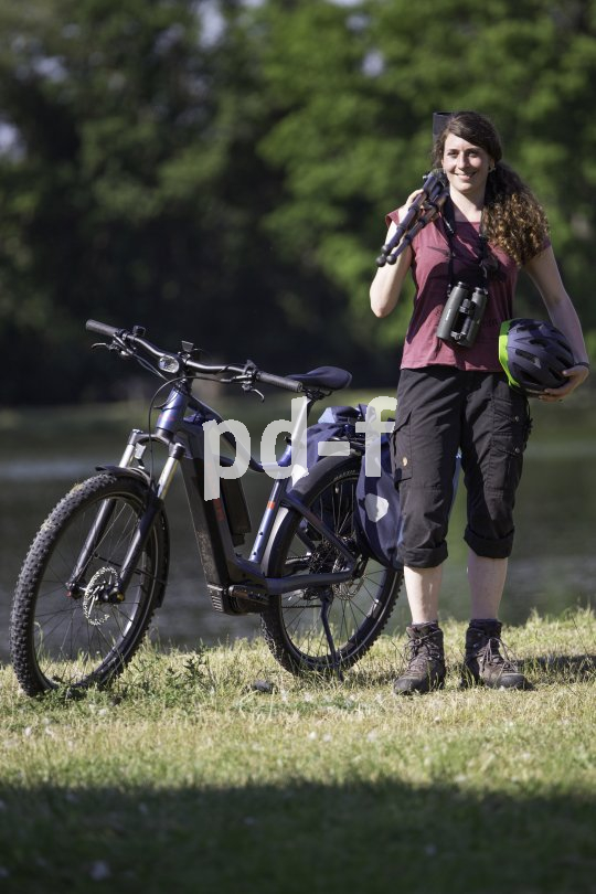 Eine Frau steht neben einem E-Bike auf einer Wiese. Auf der Schulter trägt sie ein Stativ, um den Hals hat sie ein Fernglas hängen und in der Hand einen Fahrradhelm.