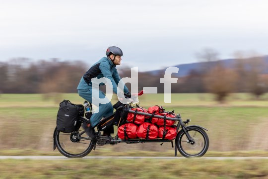 Ein Mann fährt auf einem Lastenrad mit roten Taschen auf der Ladefläche einen Radweg am Feldrand entlang.