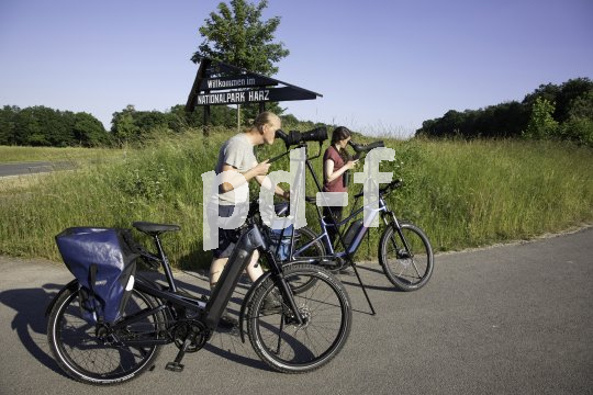 Zwei Personen stehen vor einem Schild des Nationalparks Harz und schauen in Fernrohre auf Stativen. Neben ihnen stehen E-Bikes.