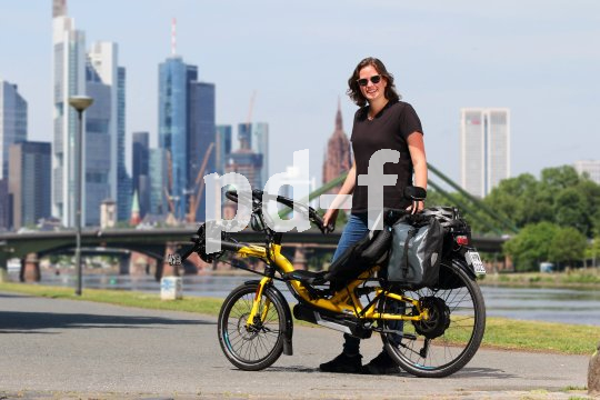 Eine Frau steht mit einem gelben Liegerad mit Versicherungskennzeichen am Gepäckträger vor der Skyline einer Großstadt.