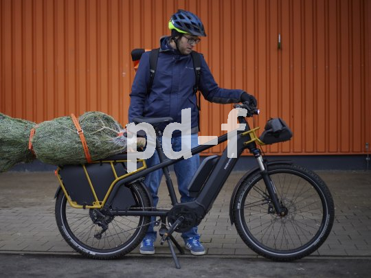 Ein Mann steht neben einem E-Bike mit einem verpackten Weihnachtbaum auf dem Gepäckträger.