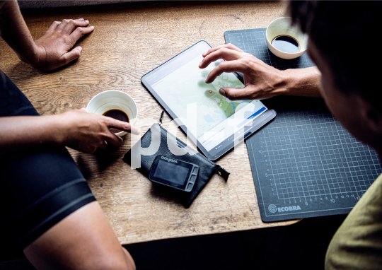 Ein Tablet liegt zwischen zwei Kaffeetassen und einem Navigationsgerät für Fahrräder auf einem Tisch. Zwei Personen beschäftigen sich mit einer Kartenanwendung darauf.