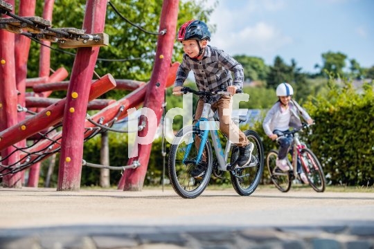 Ein Junge fährt neben einem Klettergerüst Fahrrad. Im Hintergrund folgt ein zweites Kind.
