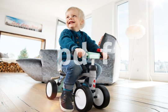 Ein lachendes Kleinkind auf einem Rutschfahrzeug in einem Wohnzimmer.