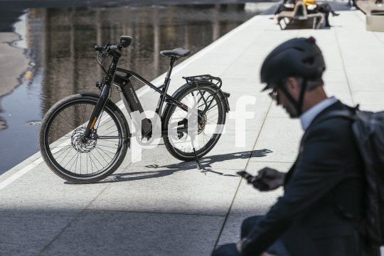 Ein E-Bike ist neben einer nassen Straße abgestellt. Im Vordergrund sitzt eine Person mit Fahrradhelm und schaut auf ihr Smartphone.