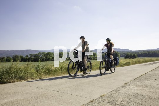Zwei Personen fahren auf E-Bikes einen betonierten Weg neben einem Feld entlang.