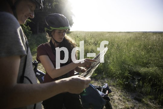 Eine Frau und ein Mann stehen vor einem Feld und betrachten eine Karte. Beide tragen Fahrradhelme.
