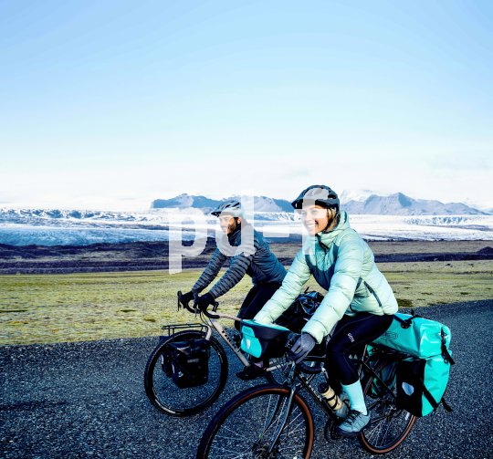 Eine Frau und ein Mann fahren lächelnd mit bepackten Fahrrädern vor einer entfernten Bergkulisse in offener Landschaft entlang.