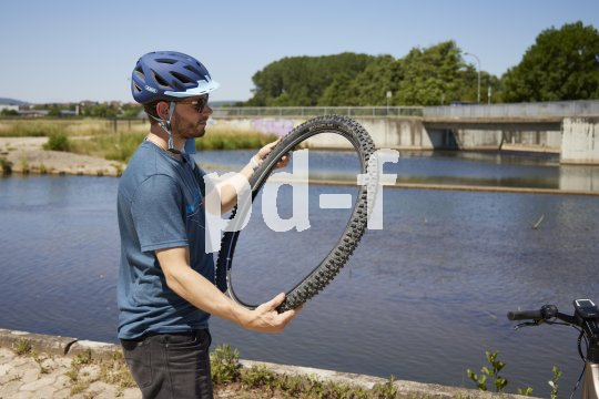 Ein Radfahrer steht vor einem Gewässer und begutachtet einen Fahrradreifen, den er mit beiden Händen vor sich hält.
