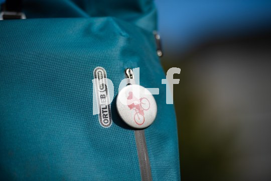 Ein runder Button mit eine:r stilisierten Radfahrer:in an einer Tasche mit dem Logo der Firma Ortlieb.