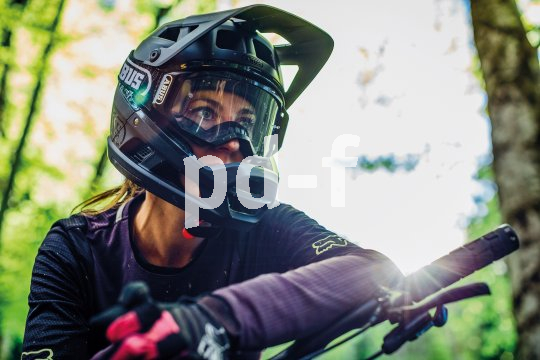 Frau mit Fullface-Helm mit Mountainbike im Wald bei Sonnenschein.