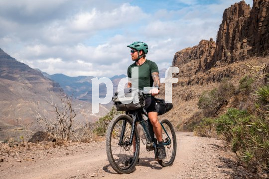 Ein Mann steht mit einem Fahrrad mit Bikepacking-Taschen auf einem Schotterweg in einer trockenen Gebirgslandschaft.