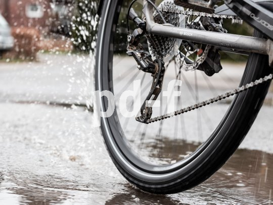 Bild des Hinterrades eines Fahrrades, dass durch eine Pfütze fährt. Vom Reifen spritzt Wasser.