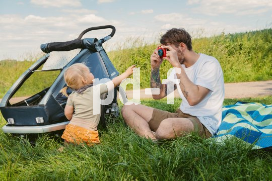 Ein Mann sitzt auf einer Wiese neben einem Fahrradanhänger und fotografiert mit einer bunten Kamera ein kleines Kind.