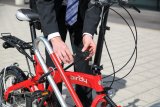 Eine Person schließt ein rotes Faltrad mit einem Fahrradschloss an einem Fahrradständer an.