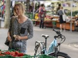 Eine Frau steht mit Fahrradhelm in der Hand an einem Gemüsestand auf einem Markt. Neben ihr steht ein E-Bike.