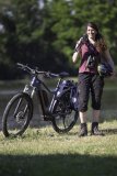 Eine Frau steht neben einem E-Bike auf einer Wiese. Auf der Schulter trägt sie ein Stativ, um den Hals hat sie ein Fernglas hängen und in der Hand einen Fahrradhelm.