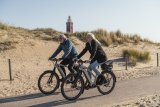 Ein Mann und eine Frau fahren auf E-Bikes einen Radweg vor einer Sanddüne entlang.