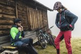 Ein Mann und eine Frau machen sich vor einer Holzhütte startklar zum Radfahren. An der Hütte lehnen zwei Mountainbikes.