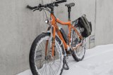 Ein orangefarbenes E-Bike mit Schnee in den Rädern und am Rahmen steht an eine Wand gelehnt.