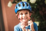 Ein lachendes Kind mit den Händen am offenen Kinnriemen eines blauen Fahrradhelmes.